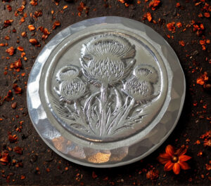 A silver round with an embossed thistle design and a hammered texture rim, resting on a dark, rustic surface scattered with dried red flower petals.