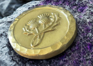 A thick, silver circular medallion featuring an embossed twin thistle design and a hand-hammered beveled edge, resting on a textured purple and grey fibrous surface.