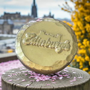 A lustrous silver art round featuring a raised silhouette of Edinburgh Castle and script, standing on a wooden post covered in pink petals with the out-of-focus city skyline in the background.